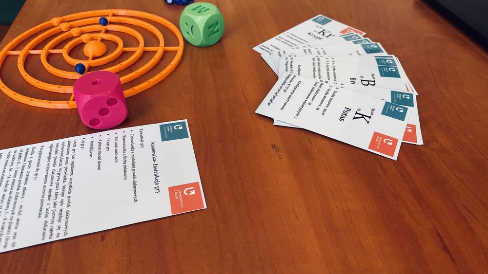 Na zdjęciu widoczne dwie kostki do gry oraz planszę w postaci okrągłych orbitali atomowych z z atomami w postaci koralików. Do zestawu dołączone są karty gry.