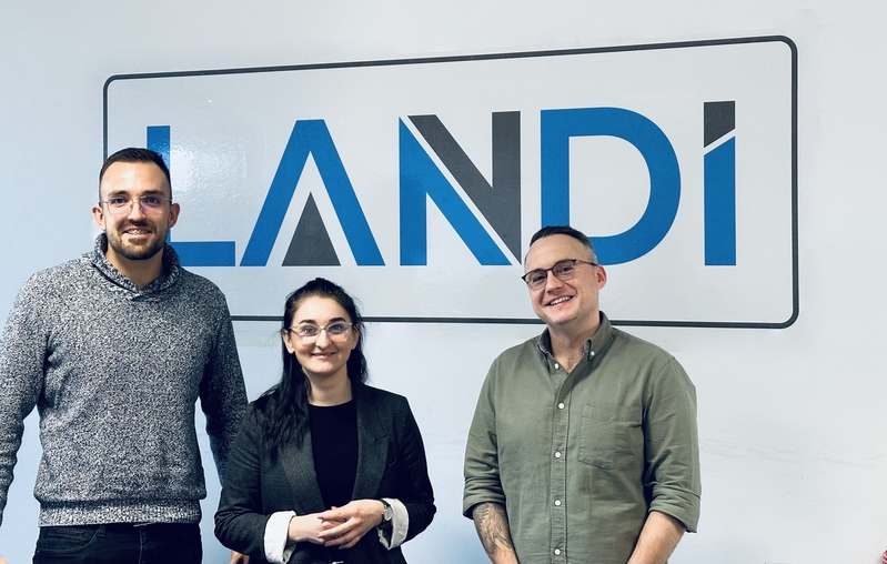 Dwóch pracowników firmy Landi oraz pracowniczka EkSocu stoją na tle dużego logotypu firmy. Od lewej: mgr Jan Fudali, dr Joanna Górniak i mgr Mateusz Piątkowski